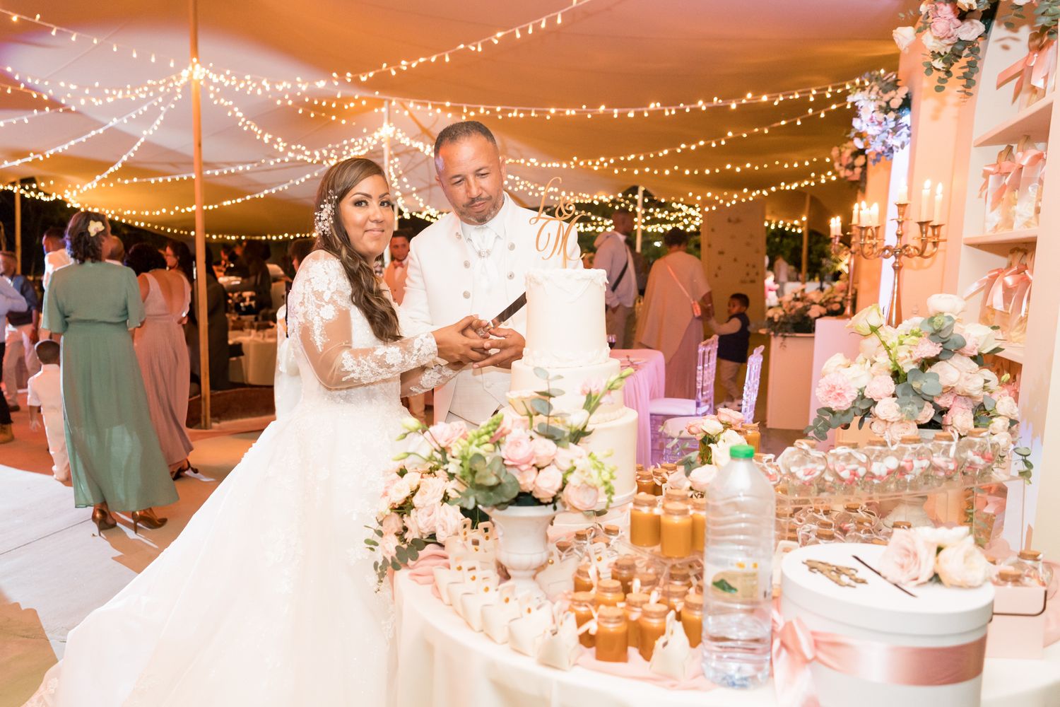 A couple cuts their wedding cake under romantic string lights at an outdoor evening reception with blush and ivory decor.