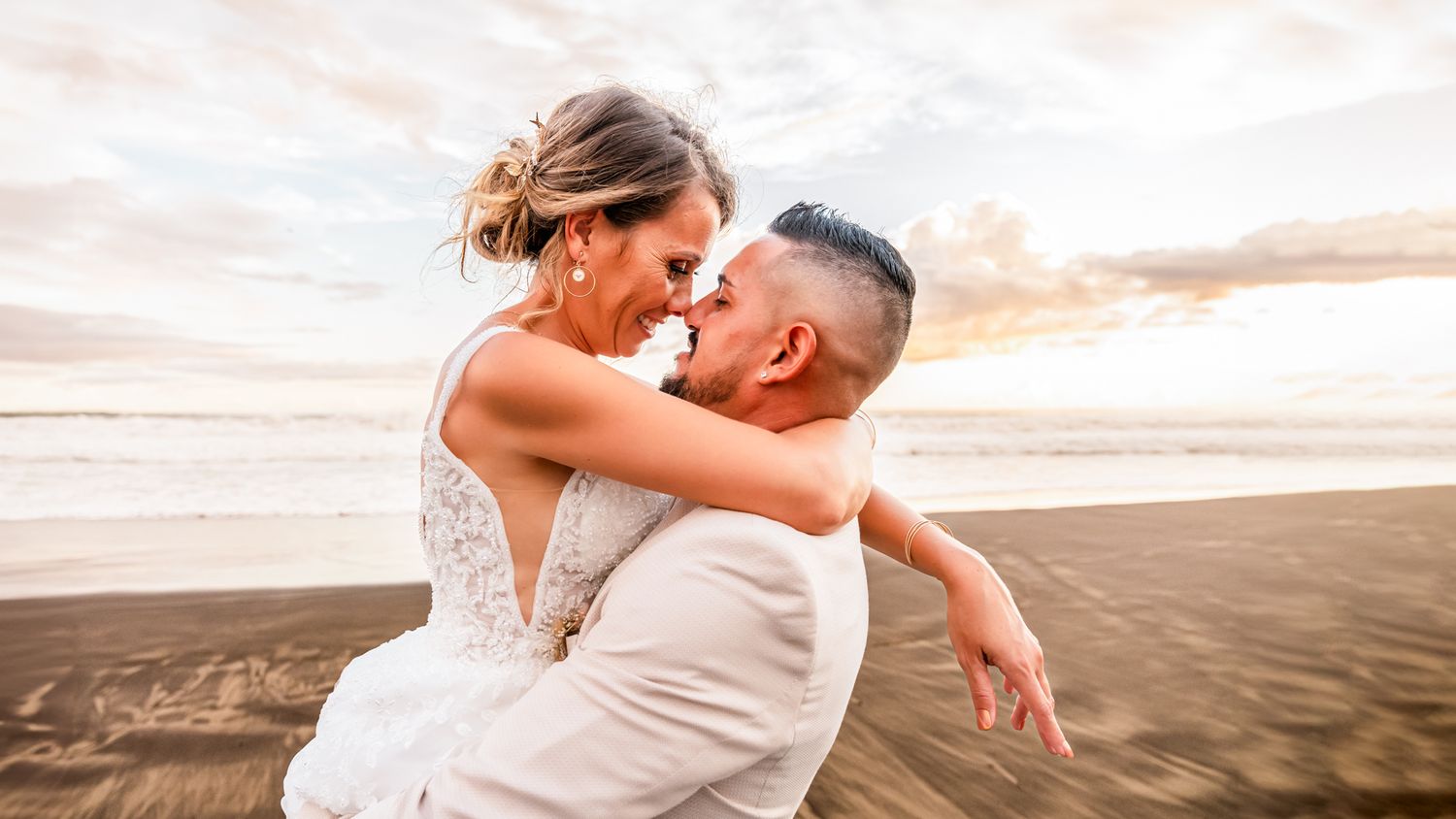 A couple in wedding attire embraces on a sandy beach at sunset with soft clouds in the background.