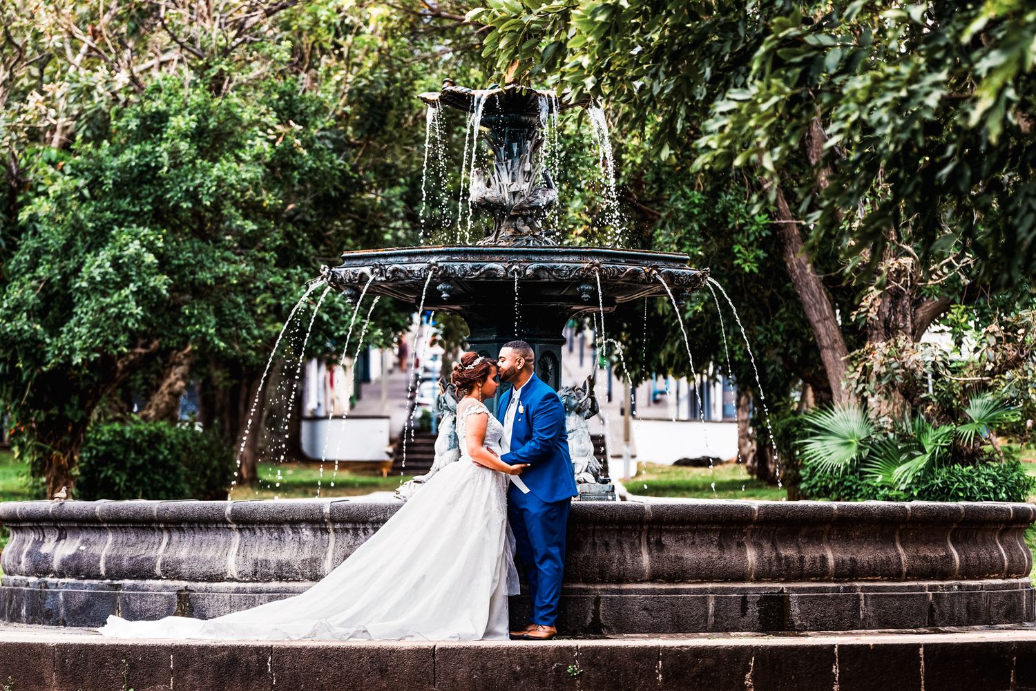A couple embraces near an ornate black metal gazebo in a lush garden setting with stone steps.