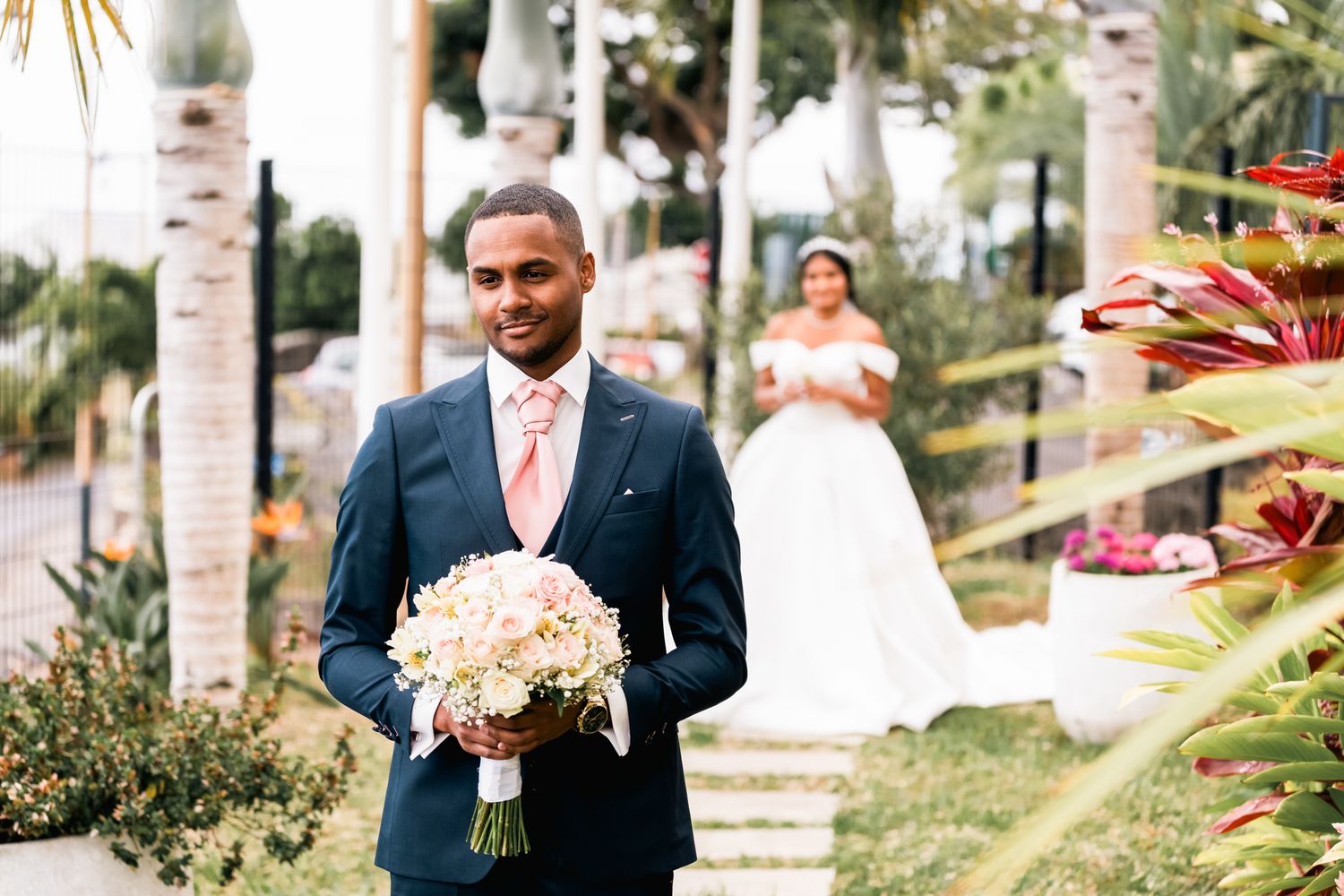 A groom in a navy suit holding a white bouquet at an outdoor wedding with palm trees and tropical landscaping.
