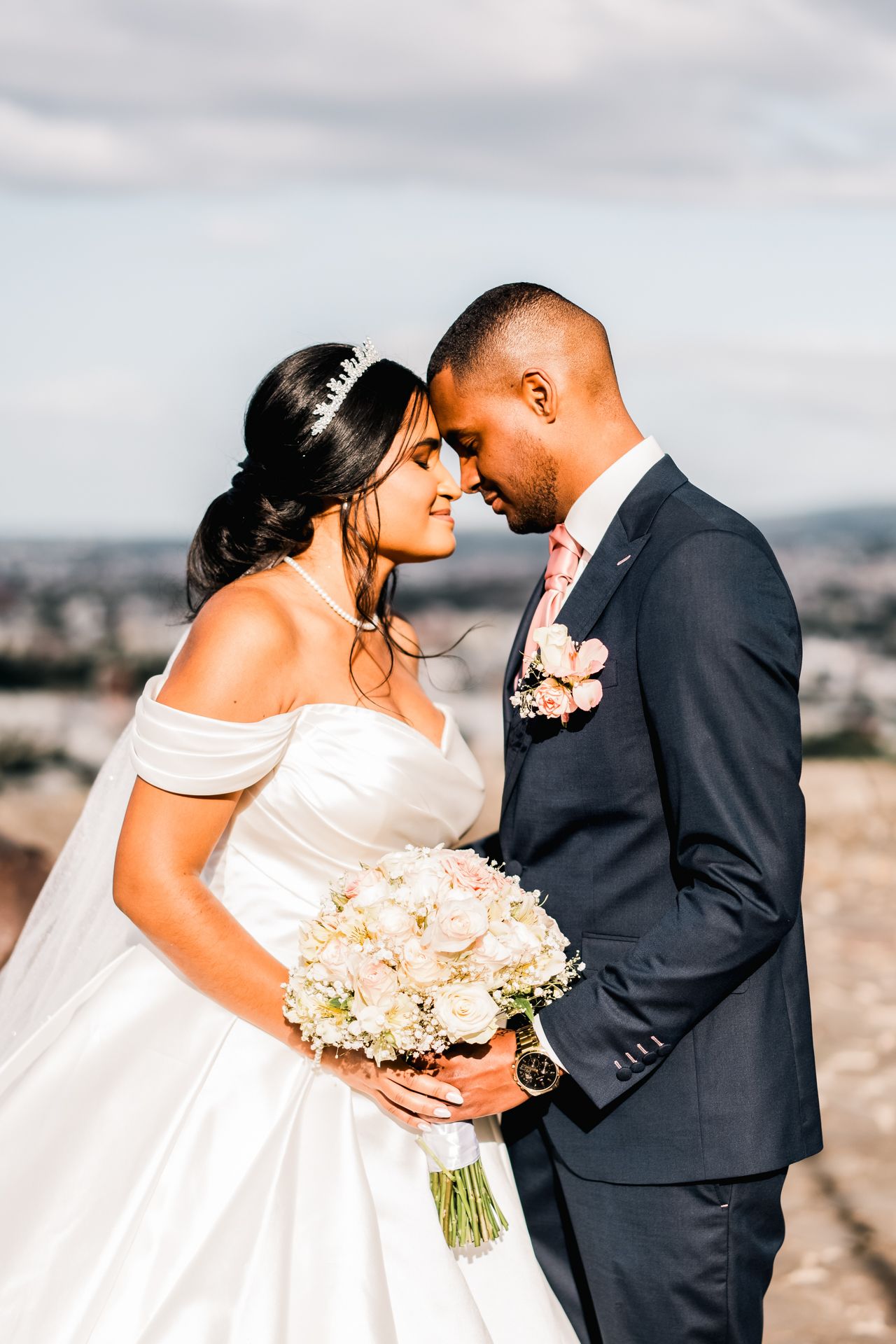 A bride in an off-shoulder wedding gown holding a white bouquet shares an intimate moment with groom in navy suit outdoors.