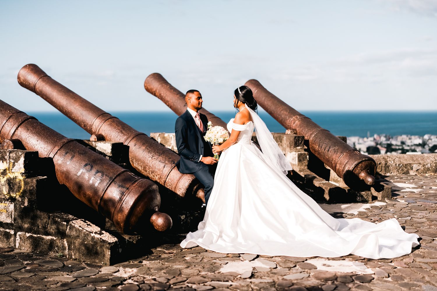 Couple in wedding attire pose romantically between historic rusty cannons overlooking an ocean view.
