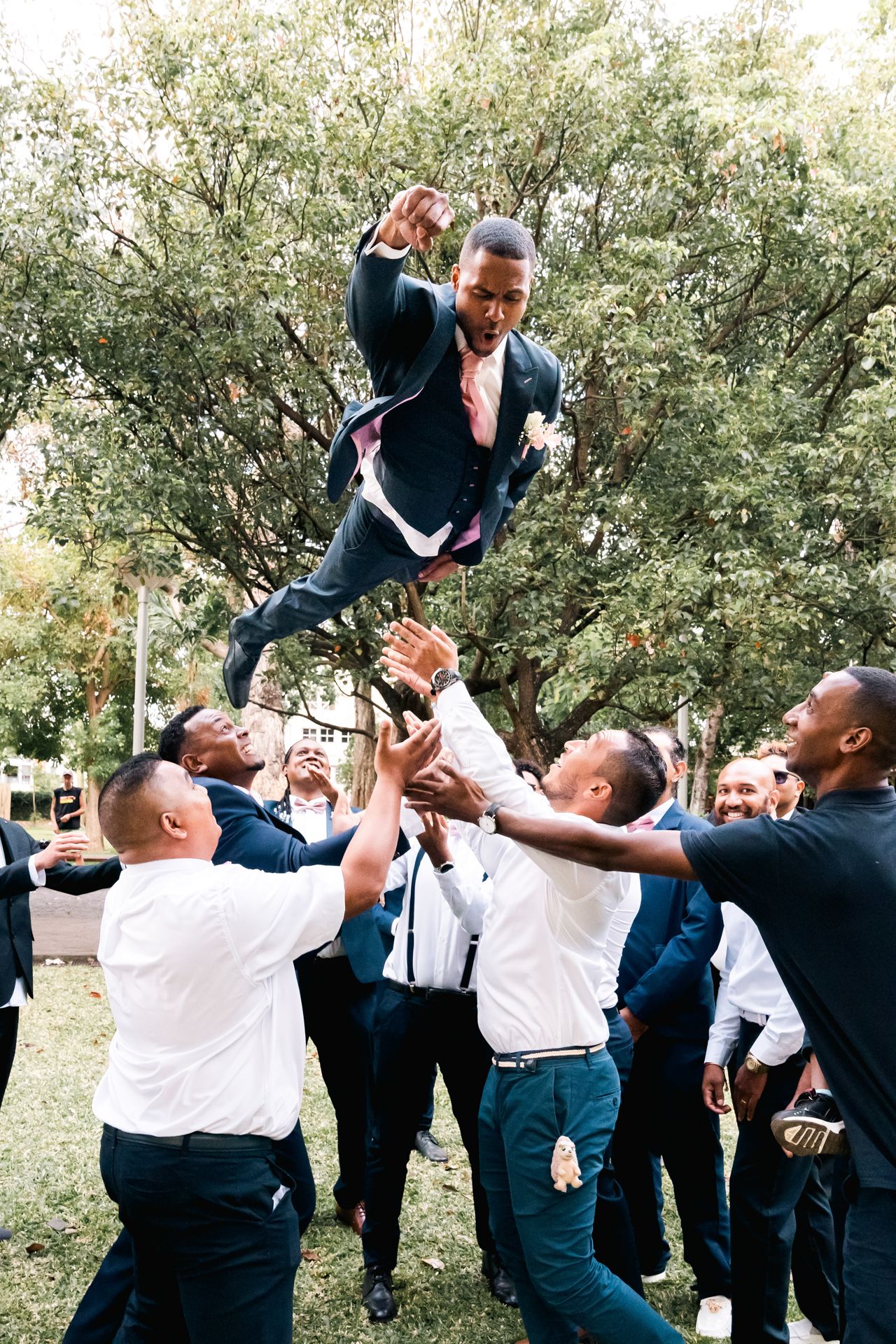 A group of groomsmen in suits lift someone up during an outdoor wedding celebration near trees.