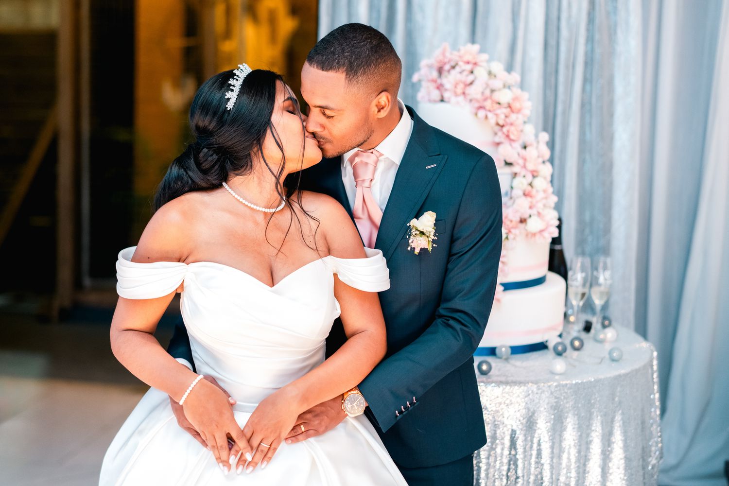 A romantic wedding moment captured with bride in off-shoulder gown and groom in navy suit sharing a kiss by their tiered cake.