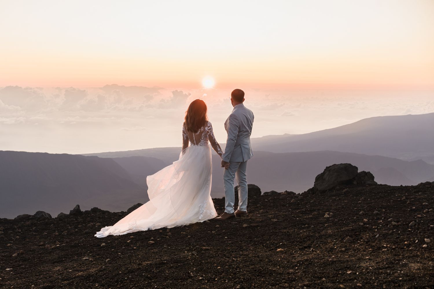Couple watching sunset from a mountain peak with flowing white dress catching the golden light against silhouetted hills.