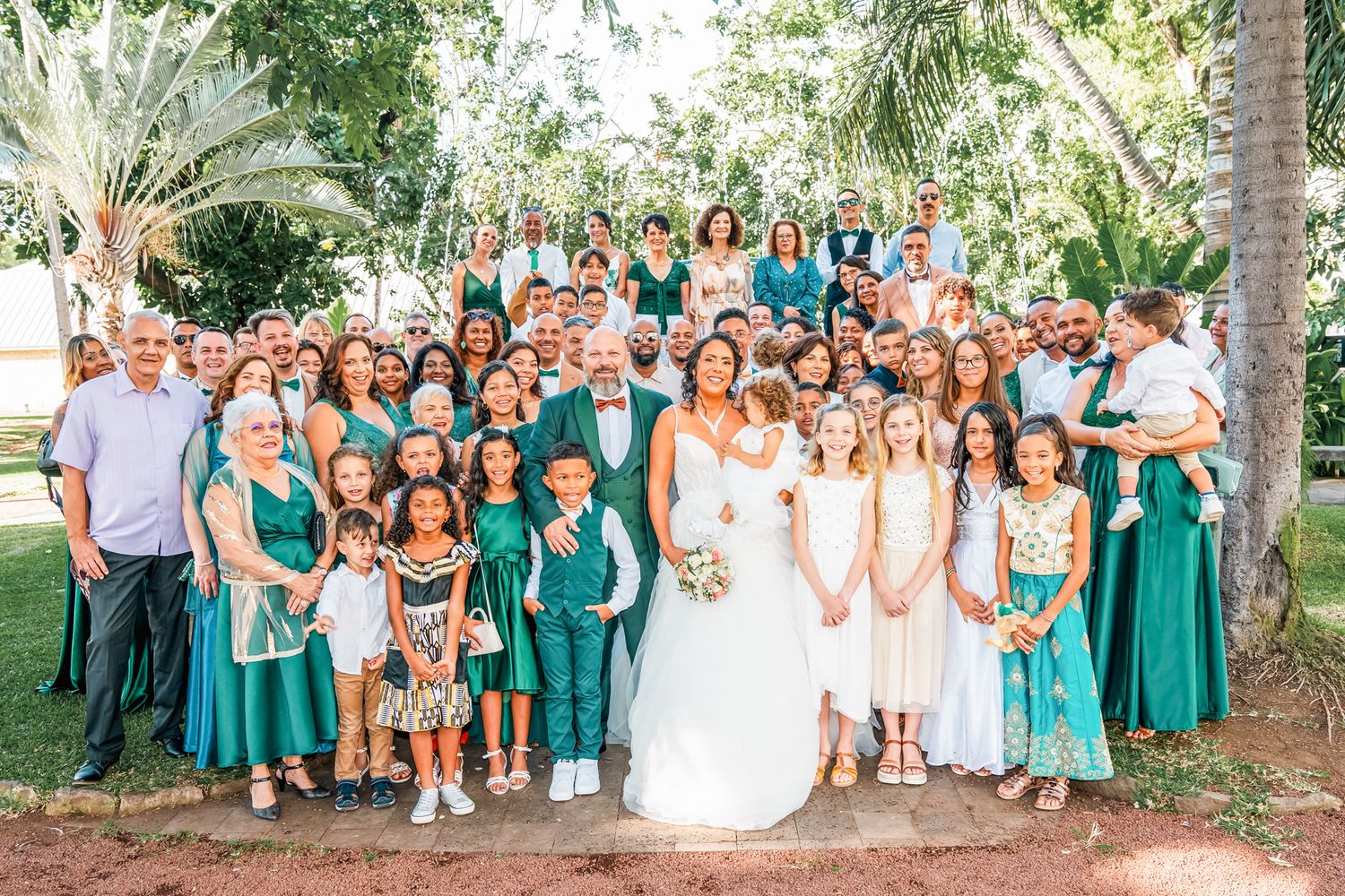 Large wedding party in green and white attire poses outdoors in a tropical garden setting.