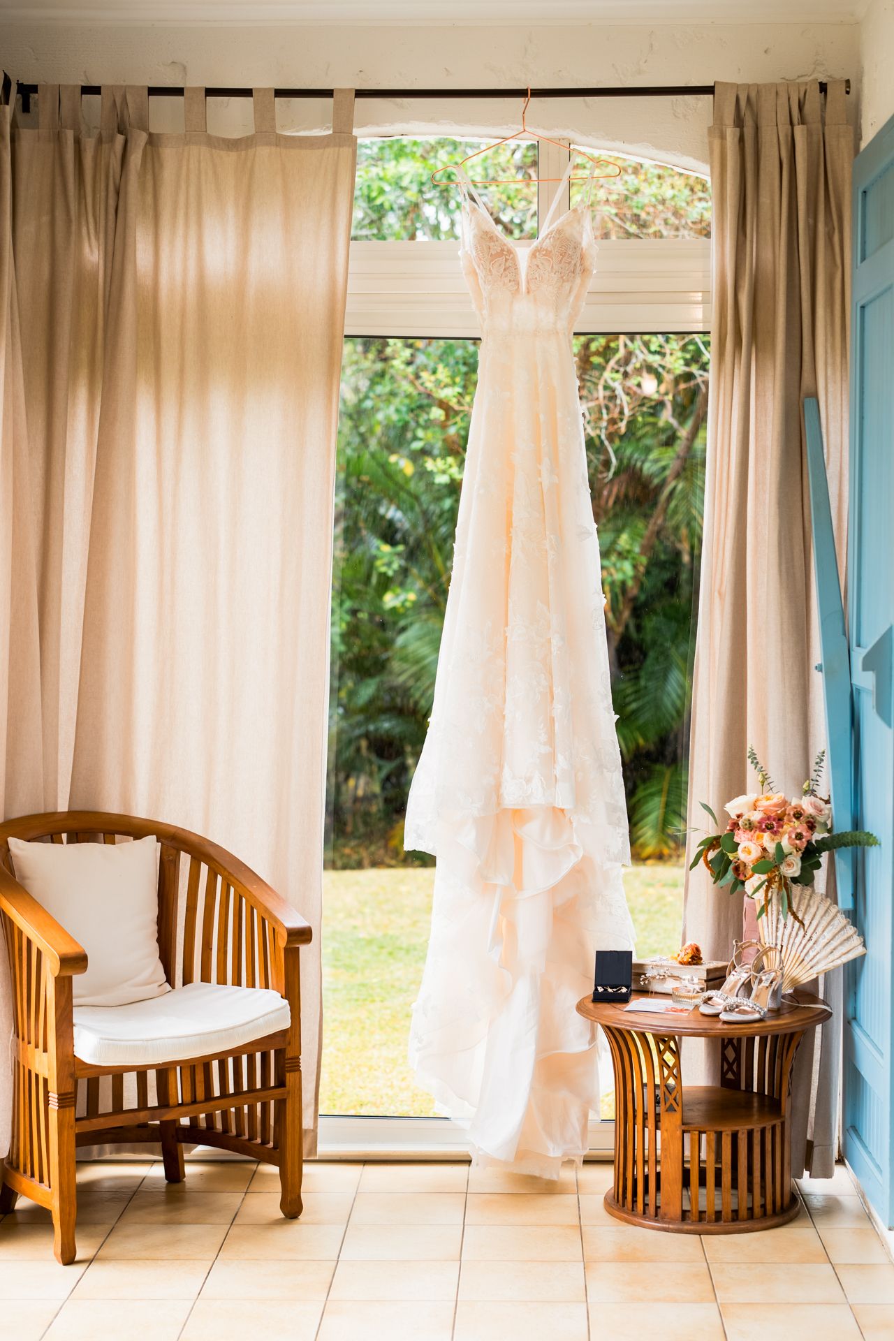 A white lace wedding dress hangs by a window with wooden furniture and pink flowers on a side table.