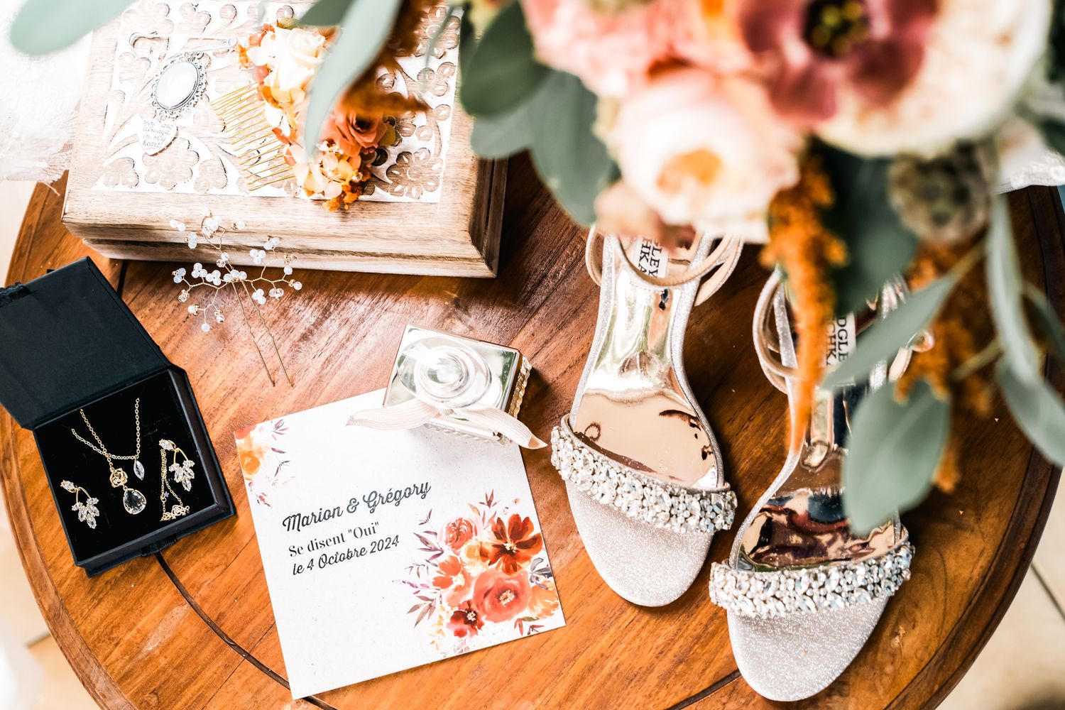 Elegant white lace bridal shoes next to wedding invitation and floral decor on wooden surface.