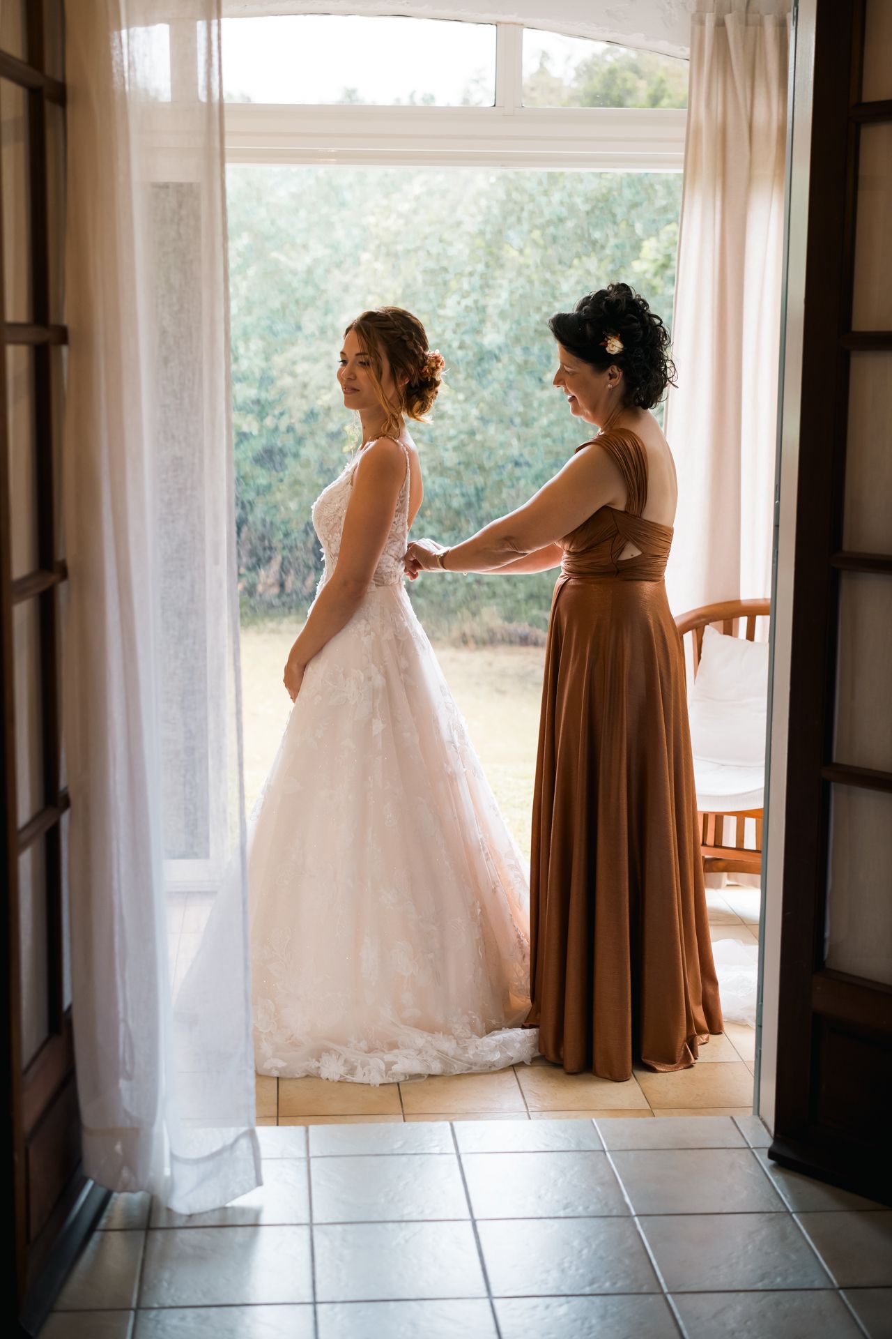 An elegant bridal scene with a white wedding dress being buttoned by someone in a brown bridesmaid gown by a sunlit window.