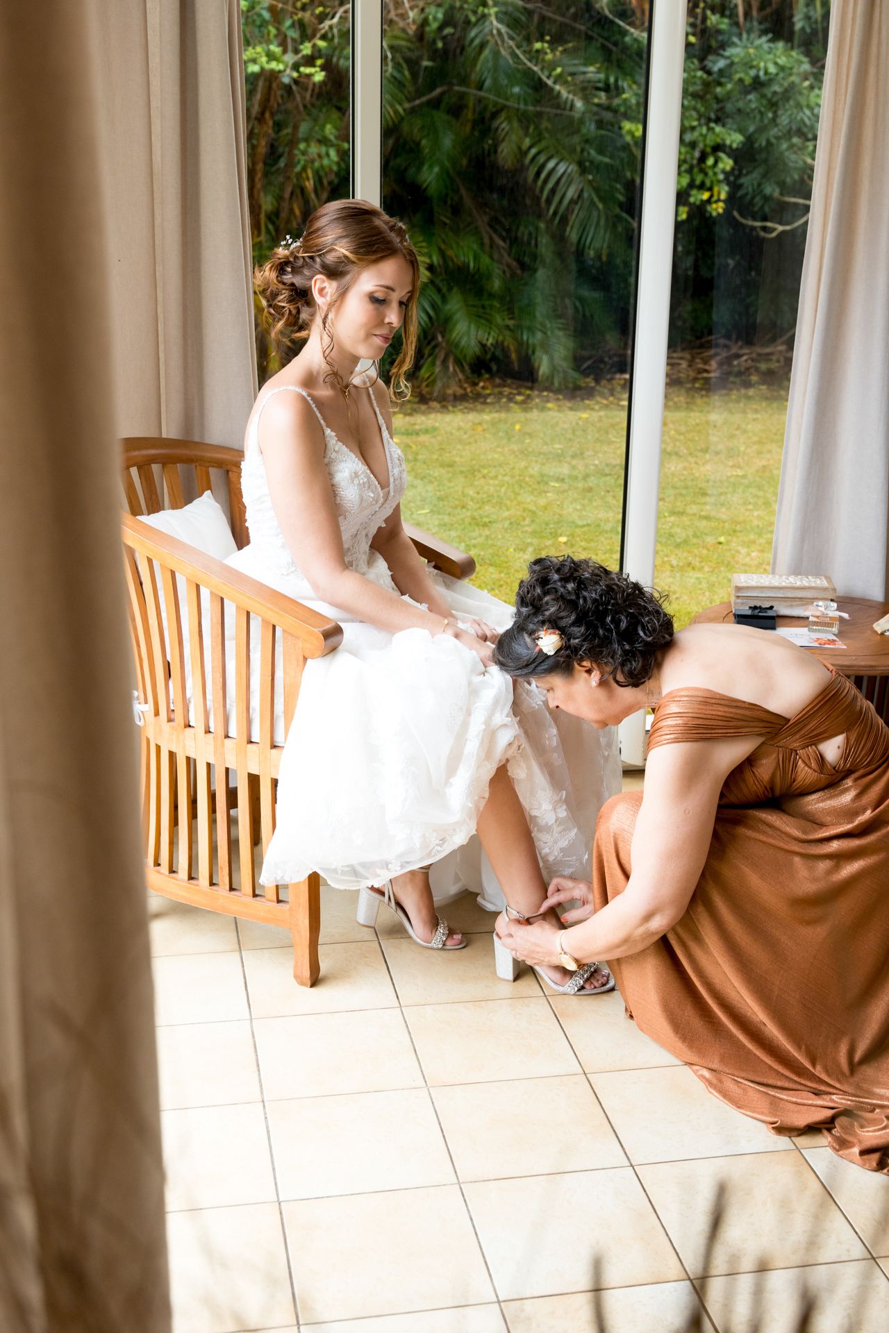 A person in a wedding dress sits on a wooden bench while someone in a brown dress helps with shoes in a sunlit room.