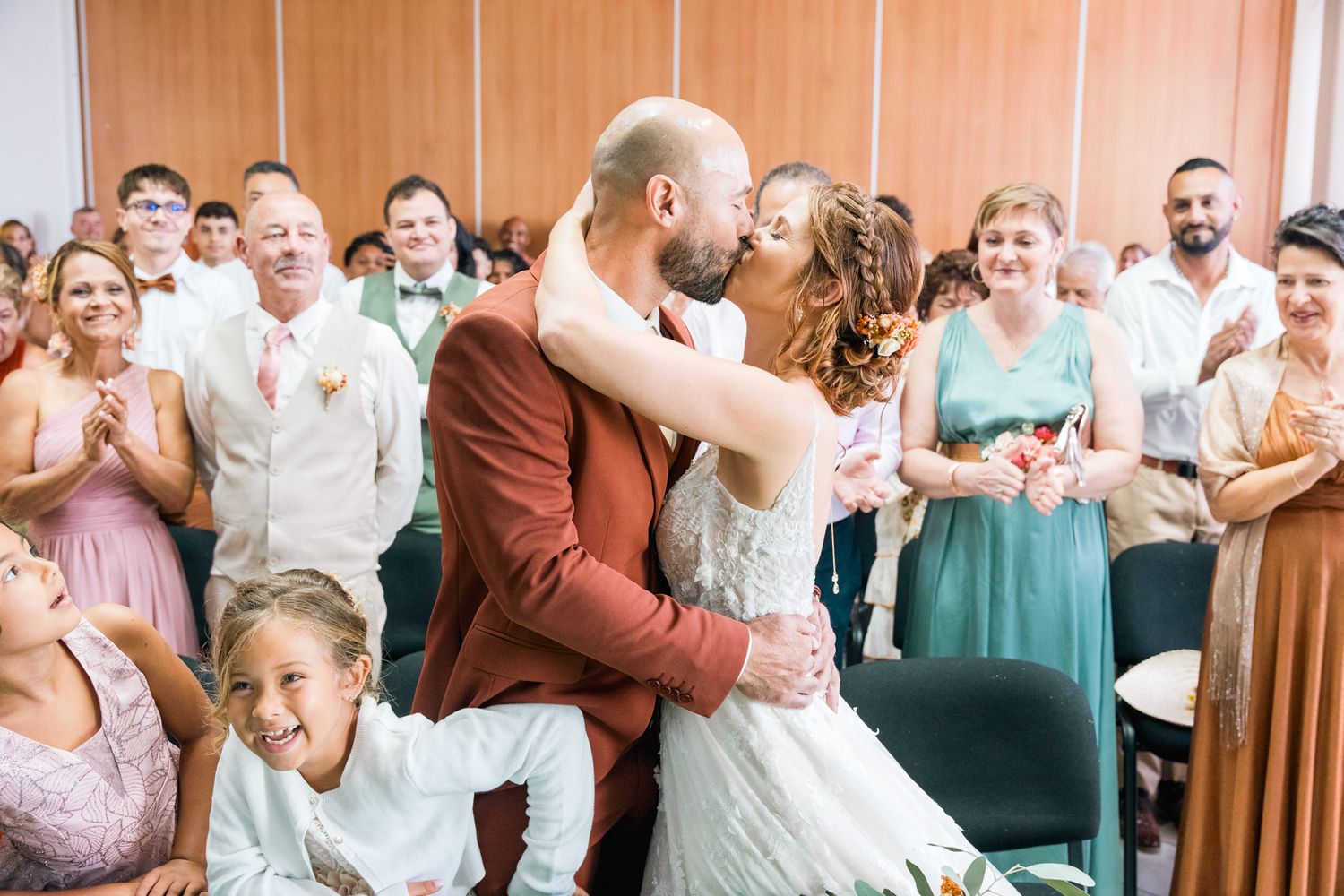 A couple shares their first kiss during a wedding ceremony while guests cheer and smile in the background.