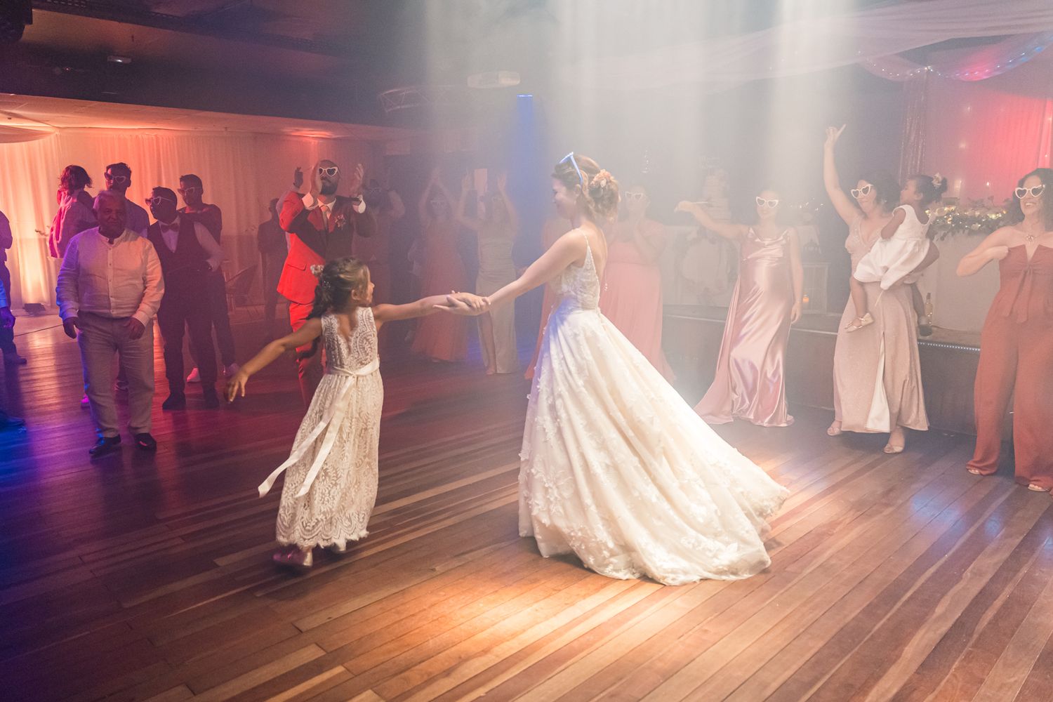 A dreamy scene at a wedding reception shows graceful dancing on a wooden floor under soft pink lighting effects.