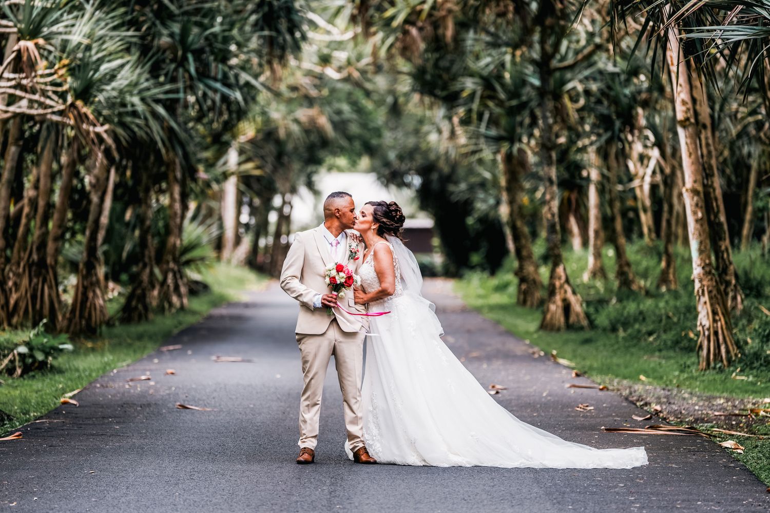 A couple in wedding attire stands on a tree-lined path with a flowing white dress and bouquet.