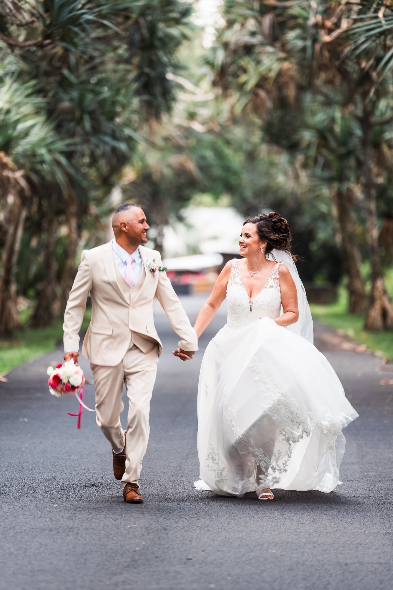 Newlyweds walk hand in hand and laugh together on a palm tree-lined road in tropical wedding attire.