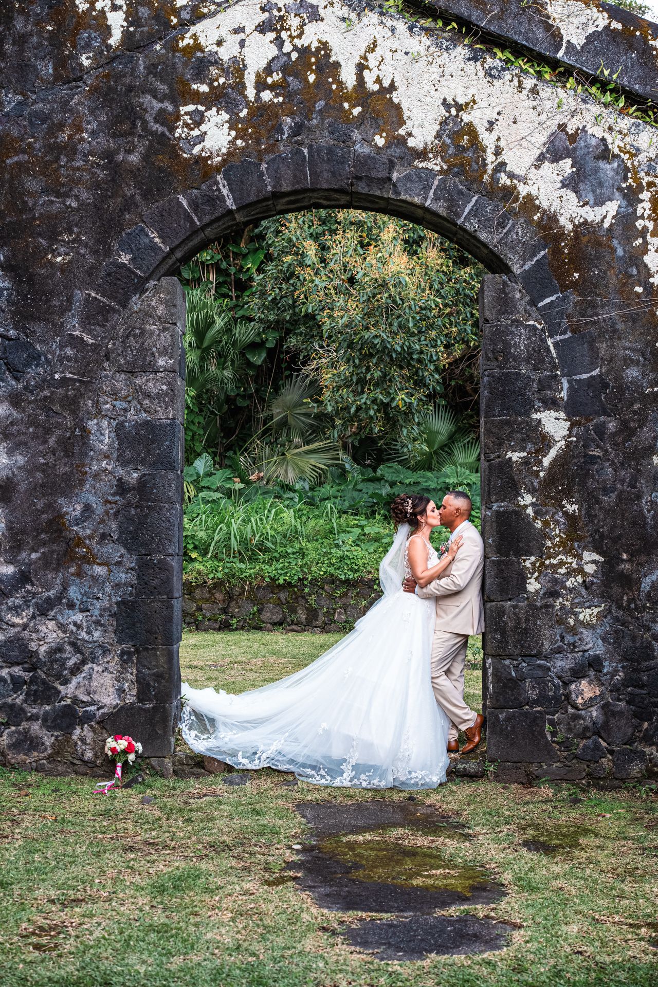 A romantic stone archway frames a wedding couple in a lush garden setting with a flowing white dress.