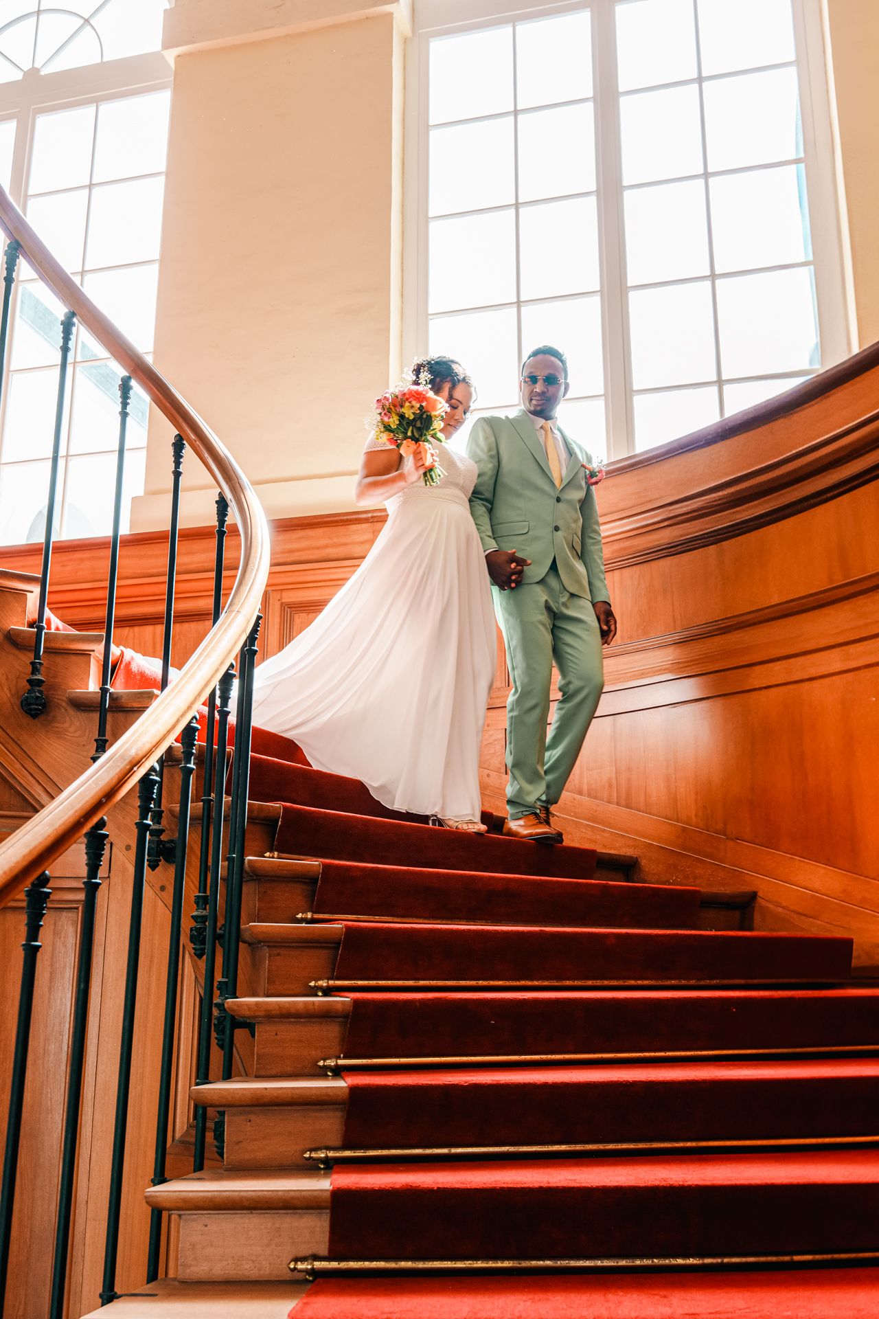 A couple in wedding attire descends an elegant staircase with red carpet and wooden banister in a sunlit venue.