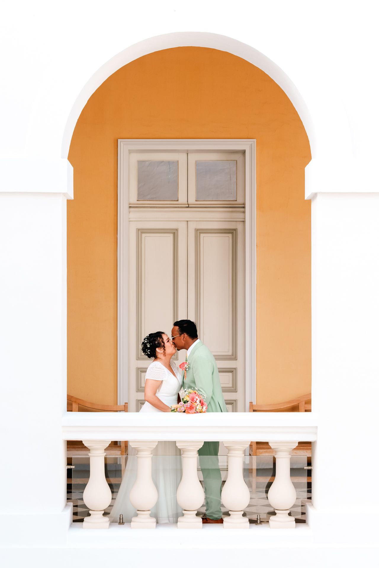 Romantic couple sharing a tender moment on an orange-arched balcony with white railings and a classic white door.