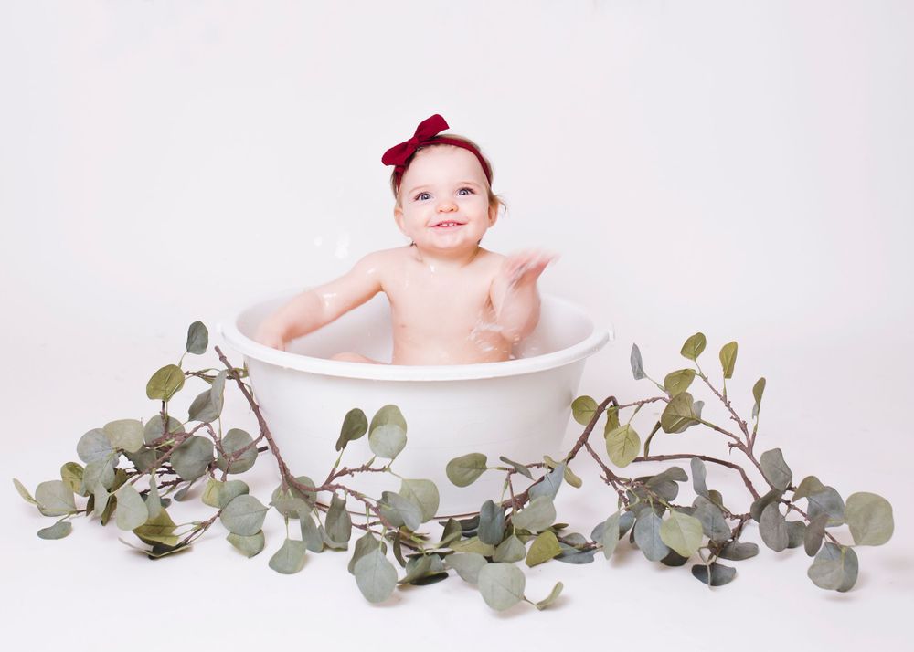 Baby sitting in white basin surrounded by decorative eucalyptus leaves and wearing a red bow.