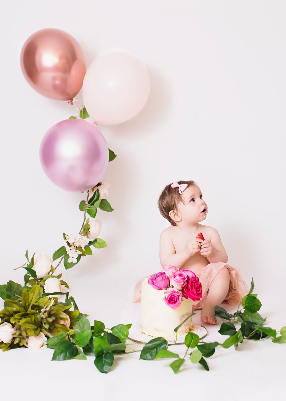 Decorative setup with pink and rose gold balloons, greenery, and floral elements against white background.