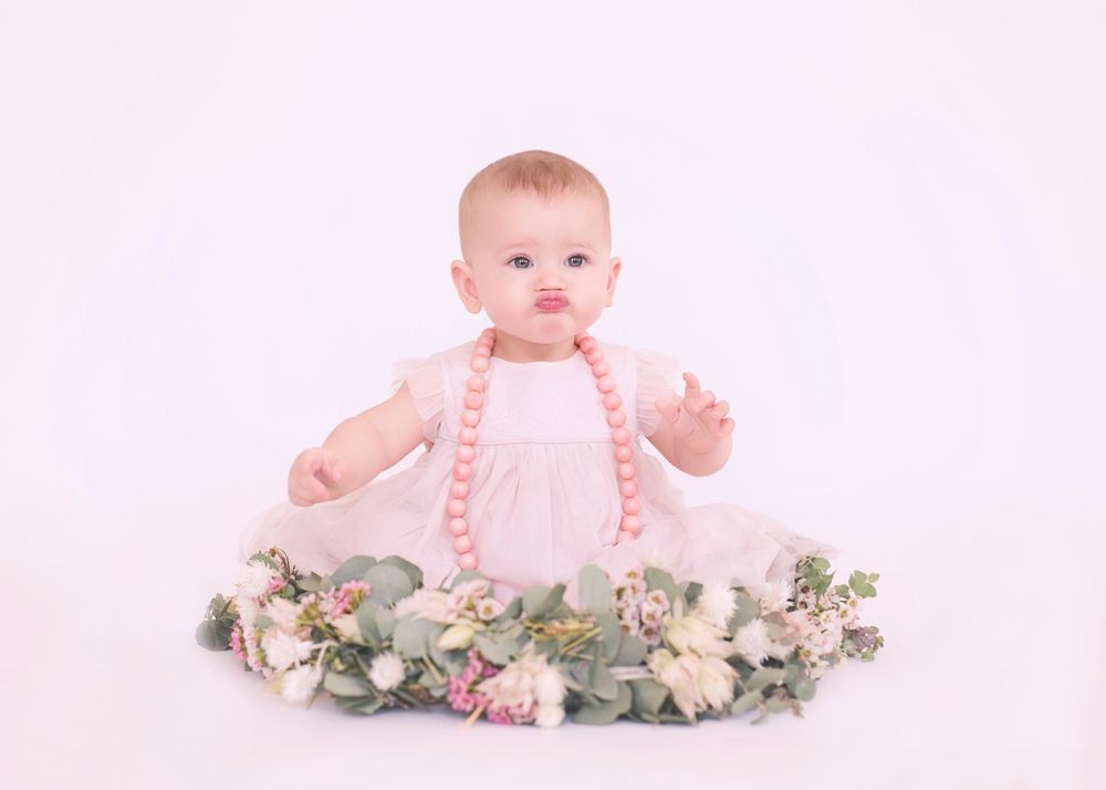 Baby in pink dress sitting among delicate floral arrangements.