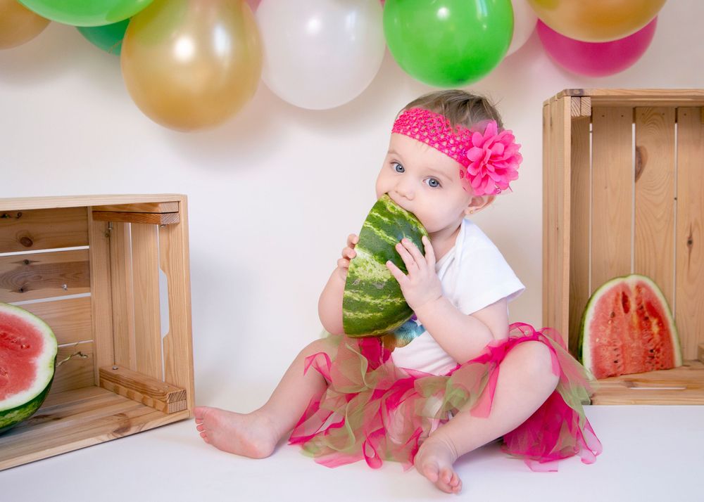 Festive setup with colorful balloons, wooden crates, and watermelon props against white background.