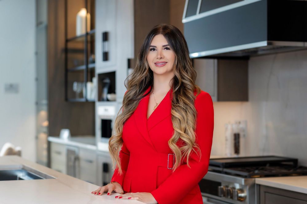 Woman in bright red blazer stands confidently in modern kitchen with stainless steel appliances and white cabinets.