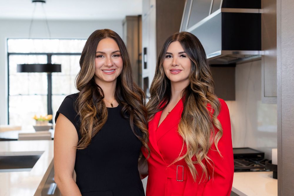 Two women in contrasting black and red outfits stand together in a modern kitchen setting.