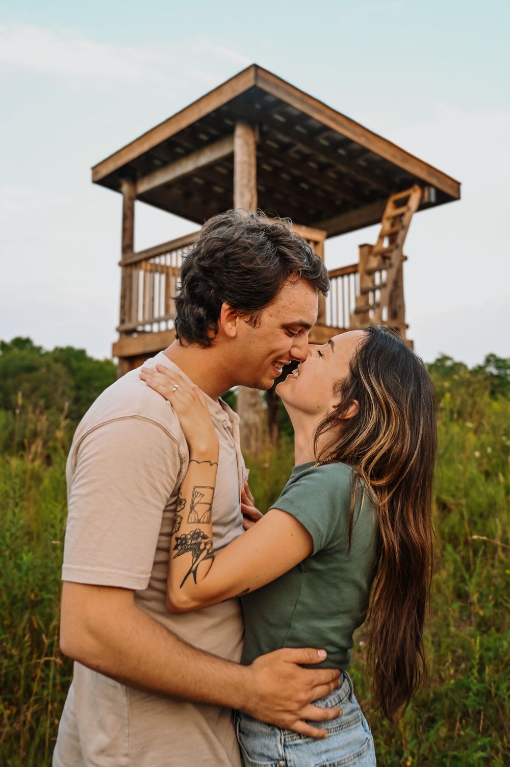 A romantic couple shares a loving embrace in front of a wooden observation tower in a nature setting during golden hour.