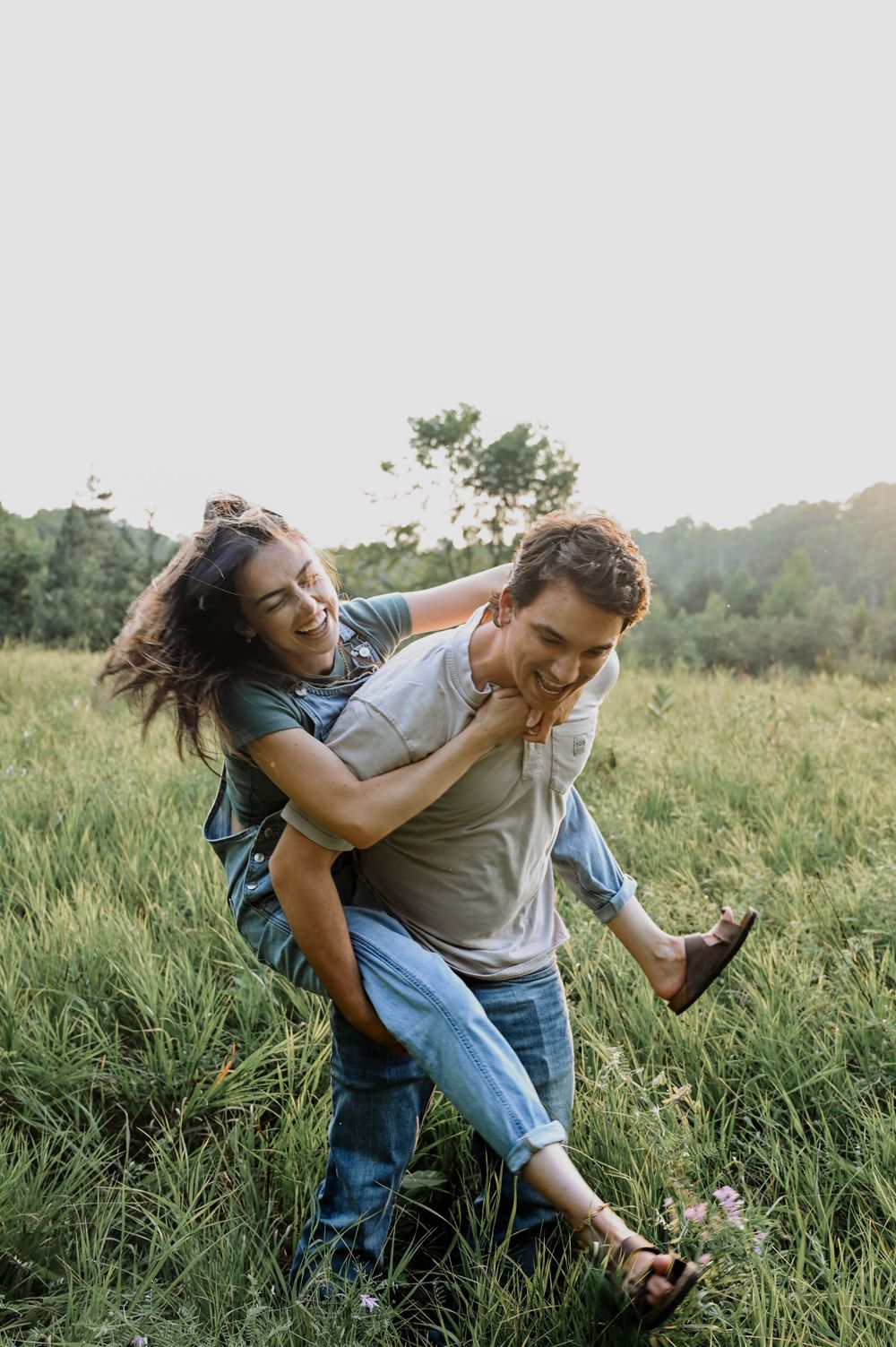 A playful couple enjoys a piggyback ride in a sunlit meadow during golden hour, surrounded by tall grass and trees.