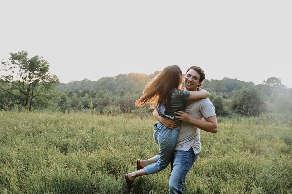 A couple embraces and spins playfully in a sunlit meadow surrounded by green trees and grass.