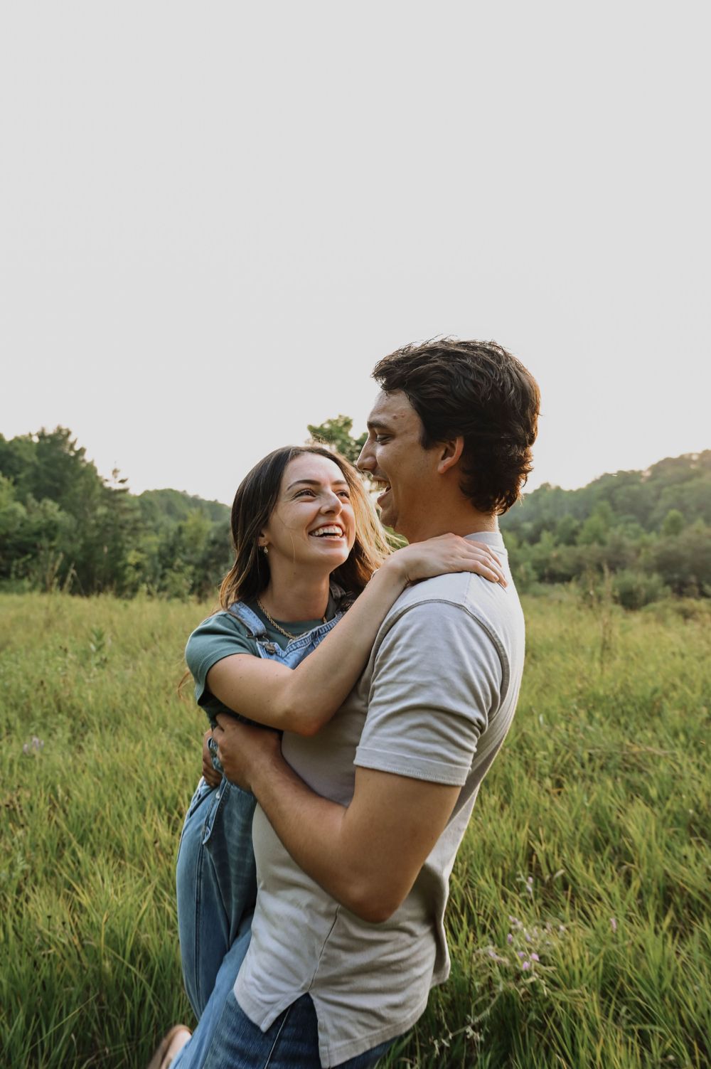 A joyful embrace between two people sharing laughter in a sunlit meadow with trees in the background.
