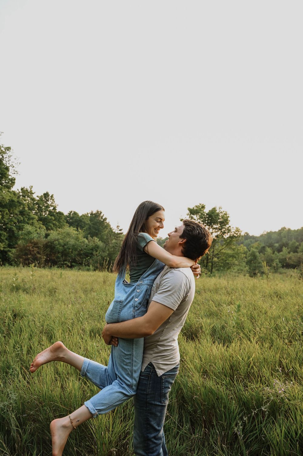 A couple embraces joyfully in a sunlit meadow, with the person in denim overalls being lifted up in a playful moment.