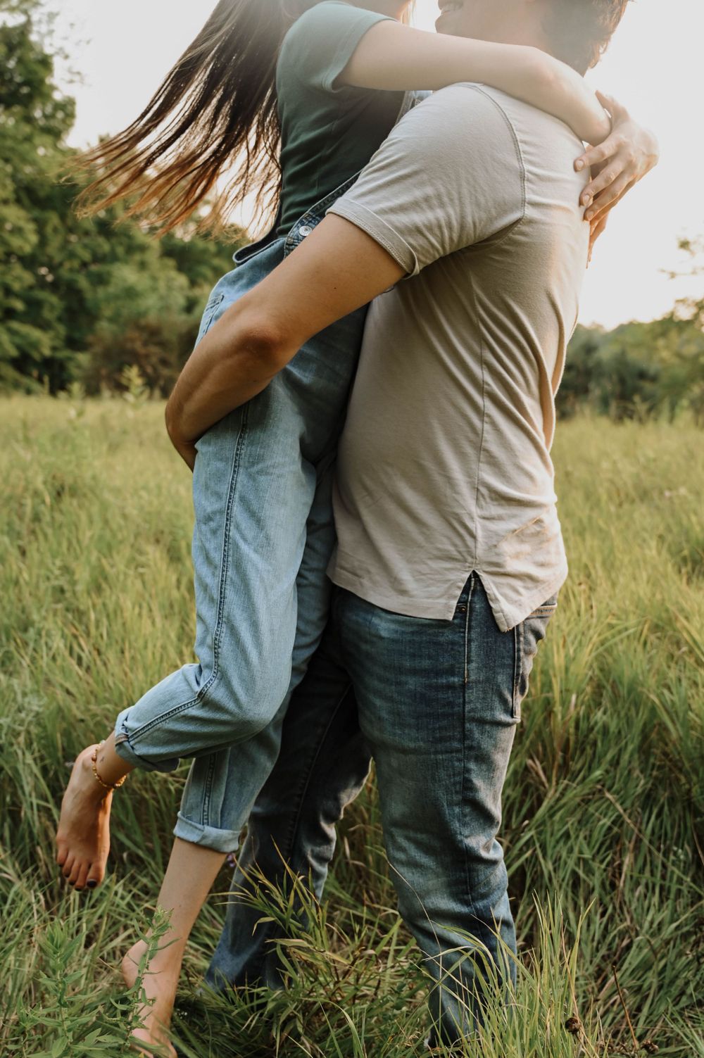 Romantic embrace in a sunlit meadow with couple standing barefoot in casual denim and t-shirts.