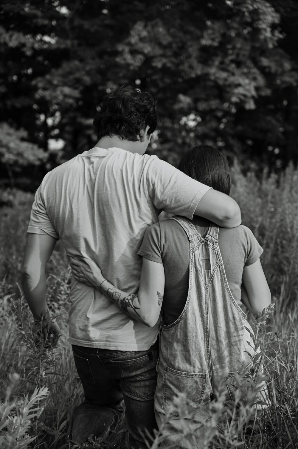 A couple embraces while walking through tall grass in a black and white romantic photograph.