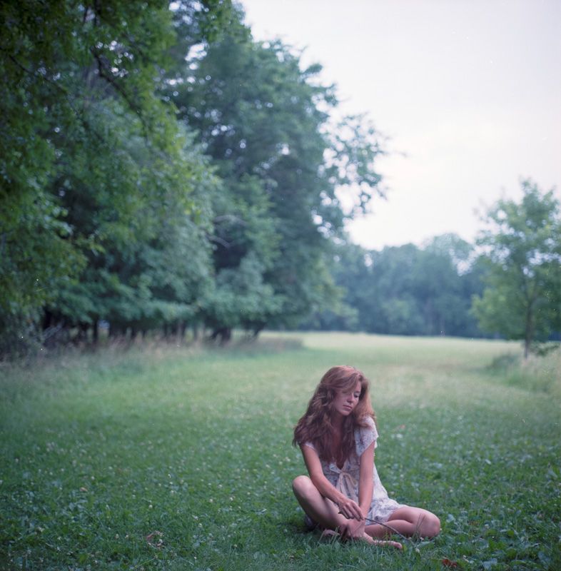 Person sitting alone in grassy field surrounded by trees.