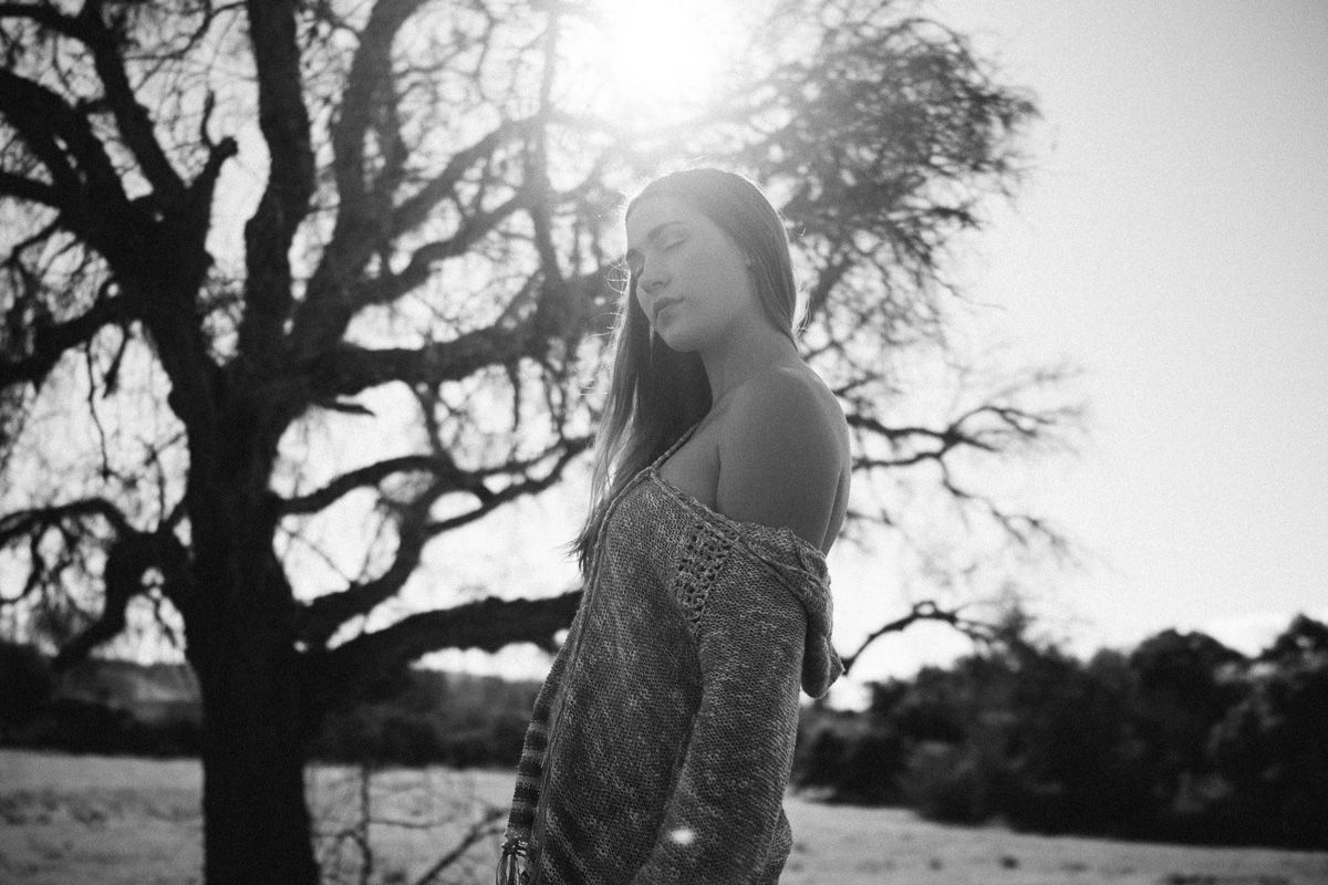 Artistic black and white portrait of a woman standing beside a silhouetted tree in soft natural light.