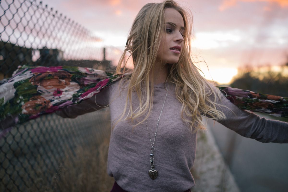 Young woman spreads her arms wide against a chain link fence during a vibrant sunset.