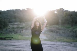Silhouetted figure in black dress poses against sunset in natural outdoor setting.