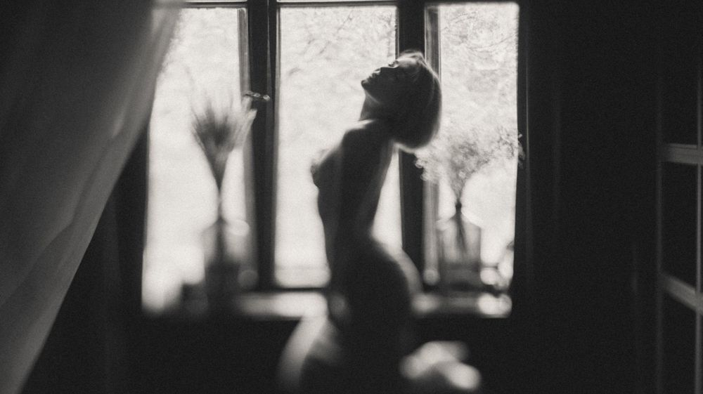 Silhouetted figure stands gracefully by a bright window in moody lighting.
