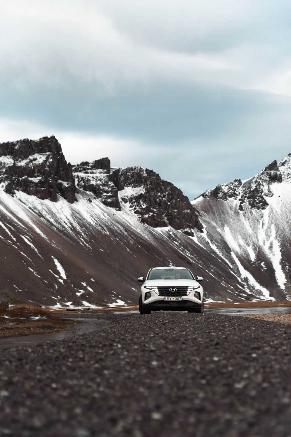 Voiture blanche conduisant sur une route de montagne avec des sommets enneigés dramatiques en arrière-plan.