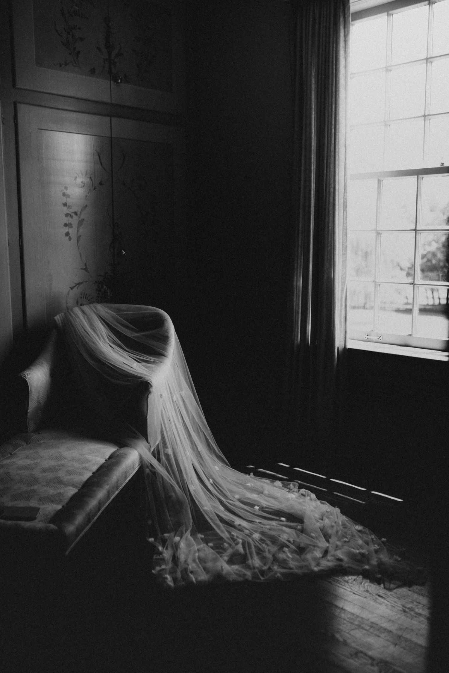 A flowing white wedding veil drapes elegantly over a chair by a bright window in a moody black and white photograph.