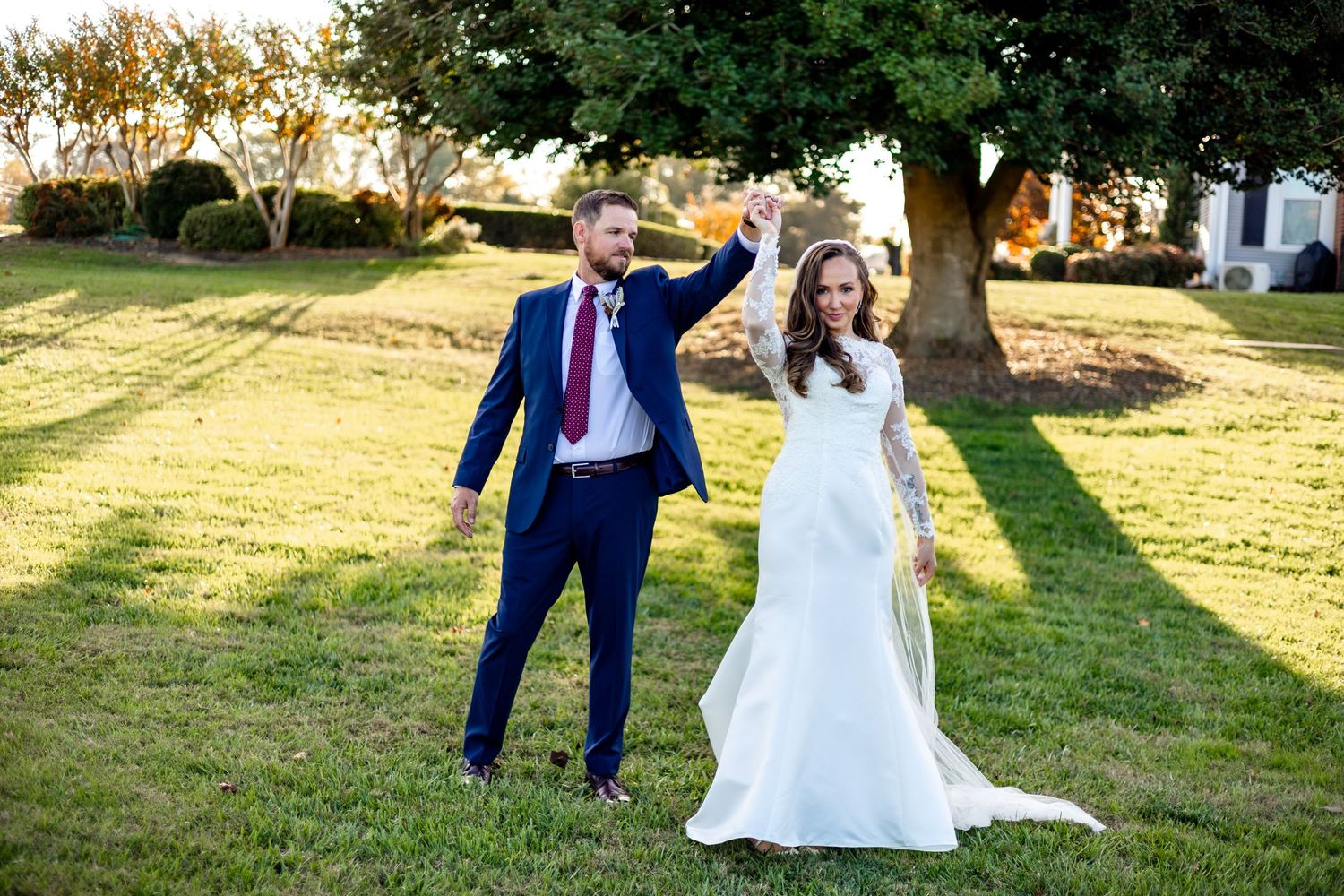 Joyful newlyweds dancing on grass under tree at sunset.
