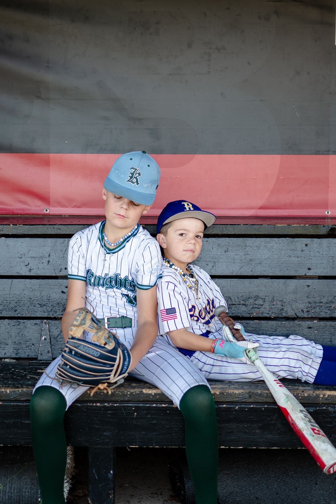Two young baseball players in different team uniforms sit together on dugout bench with their equipment.