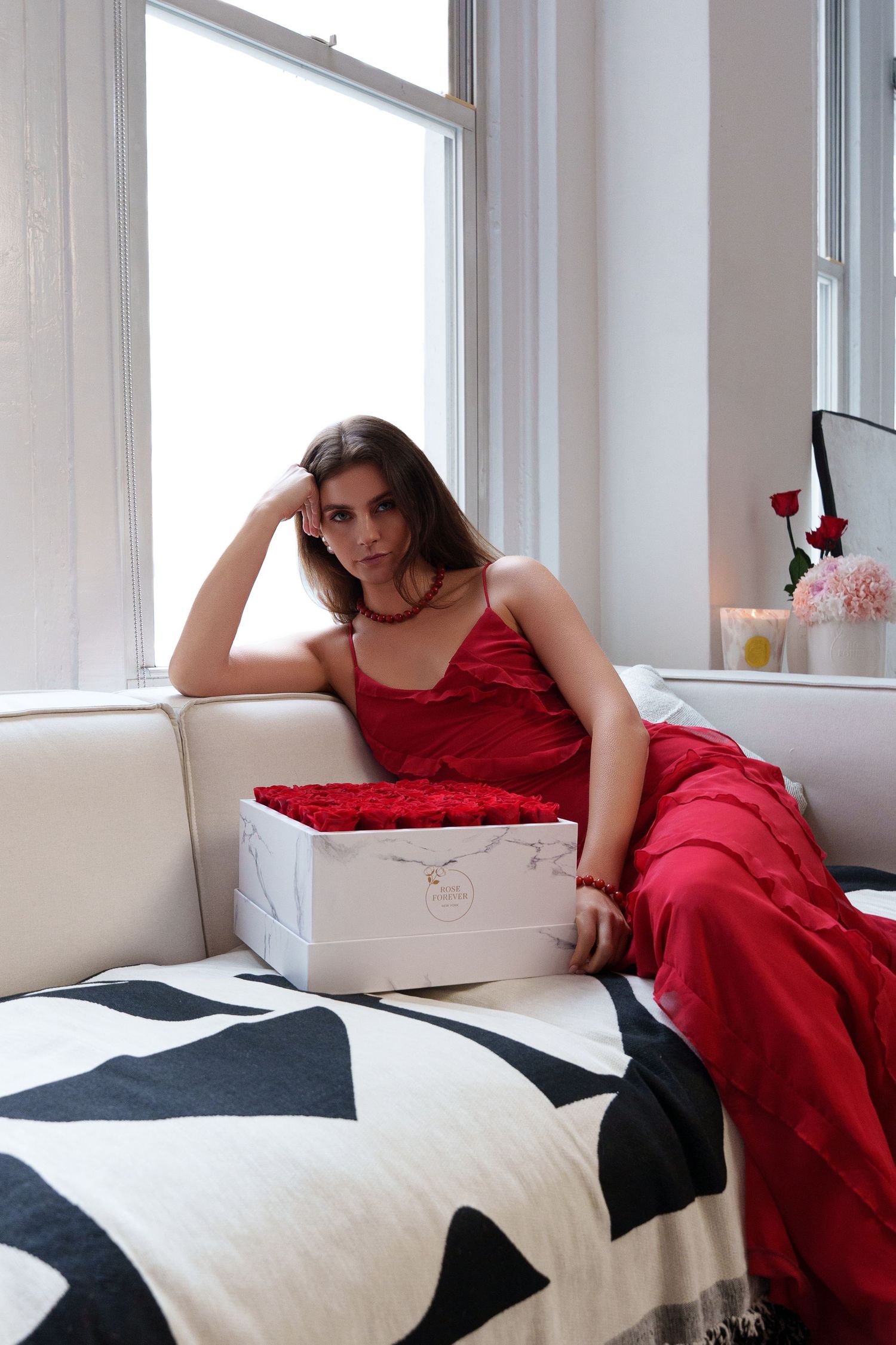 A figure in a red silk dress reclines on a white couch with geometric patterns next to a gift box near a window.