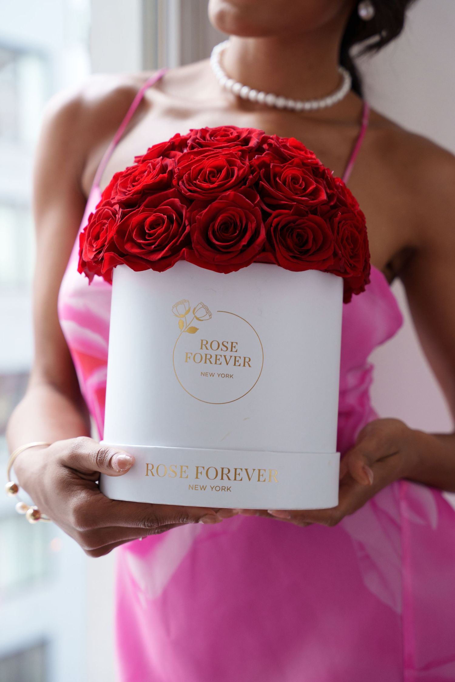 A white box filled with vibrant red roses labeled 'Rose Forever' held by someone wearing a pink dress and pearl necklace.