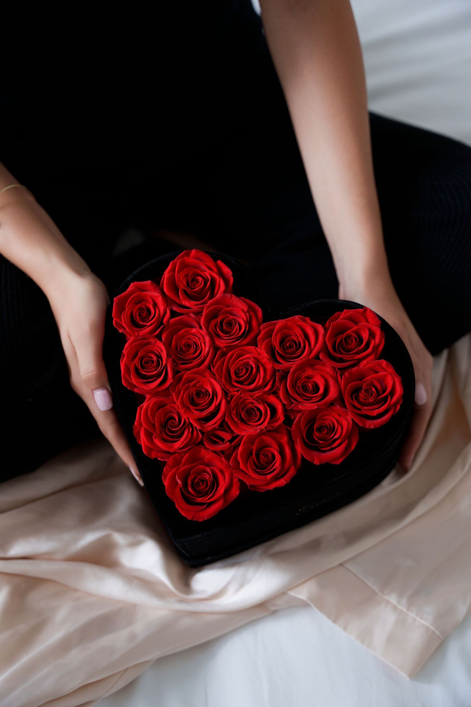 Red roses arranged in a heart shape on black velvet held by gentle hands with soft satin fabric underneath.