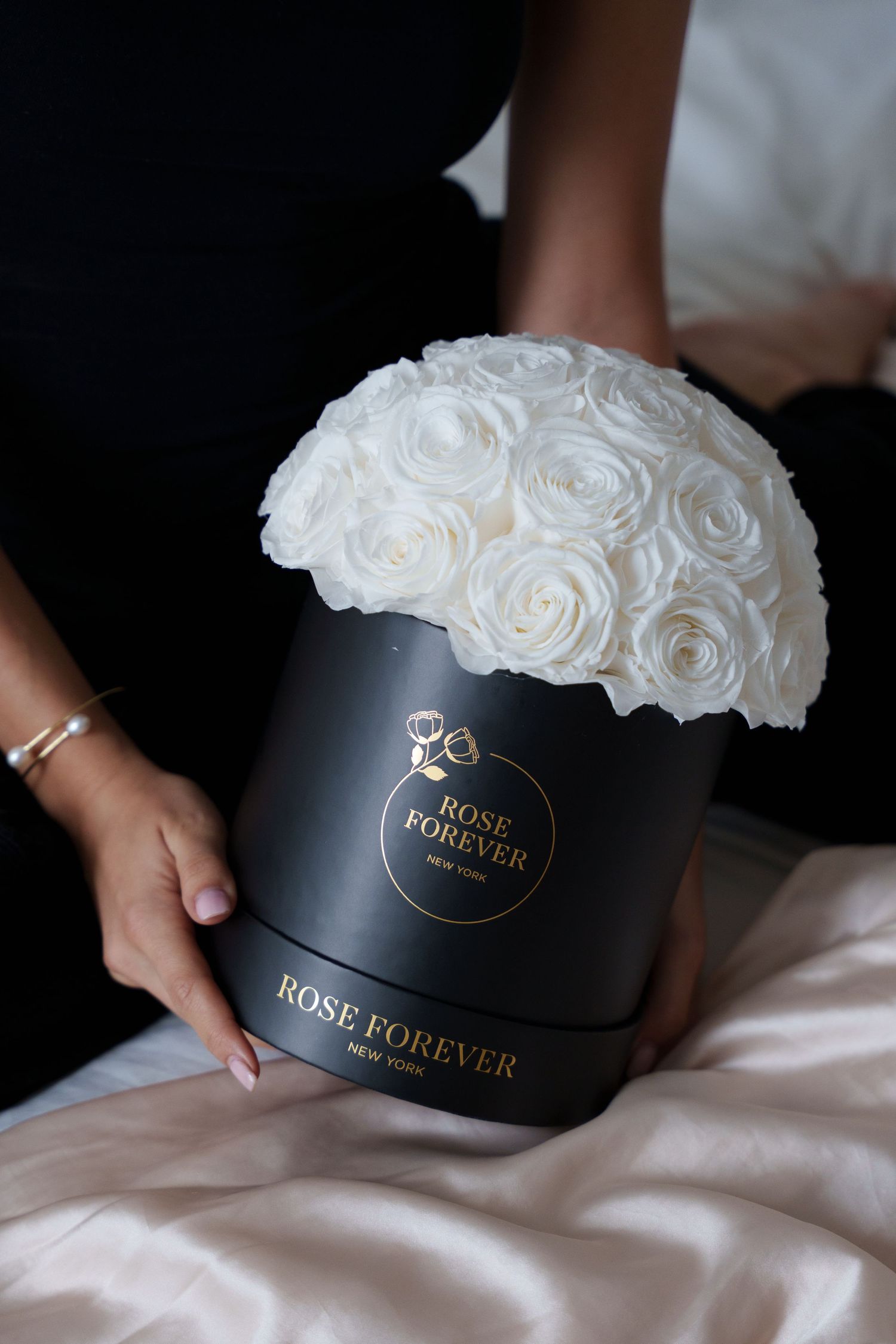 Elegant white roses arranged in a black cylindrical Rose Forever box on white fabric.