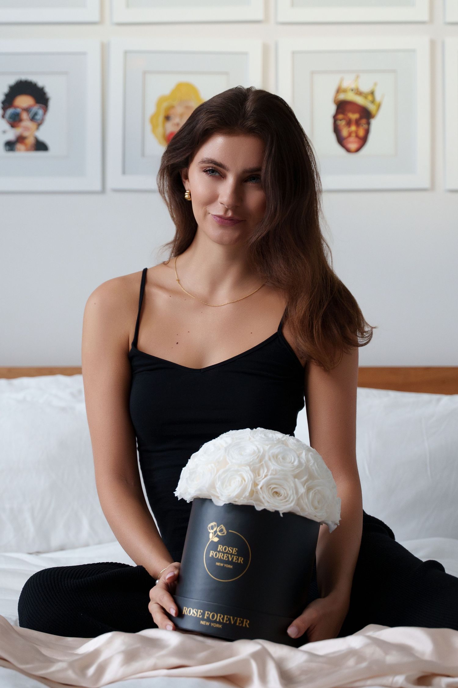 Someone in a black tank top holds an elegant white rose bouquet in a black box against a white wall with artistic portraits.