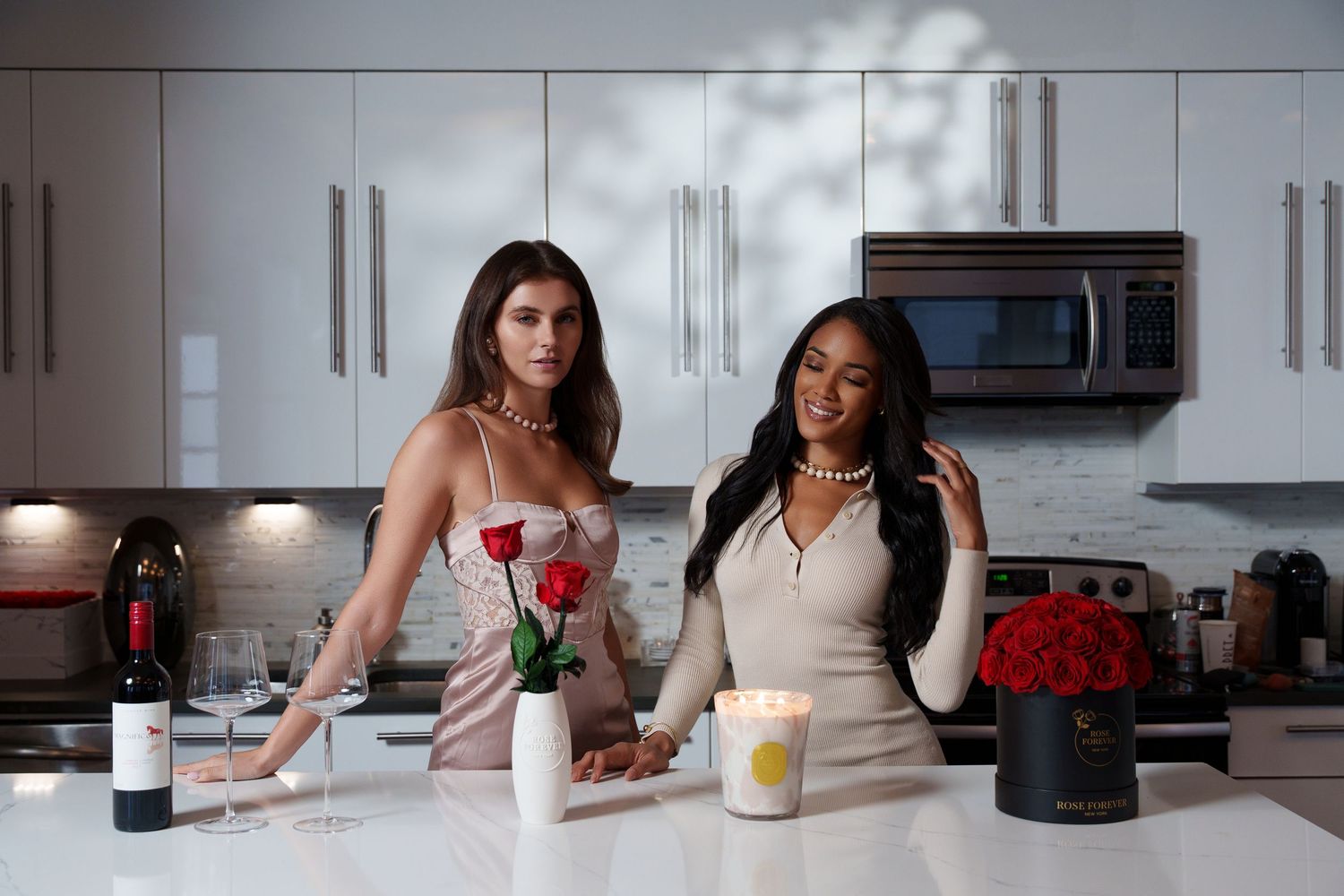 Two friends stand at a modern kitchen counter with drinks and flowers in a bright, stylish setting.