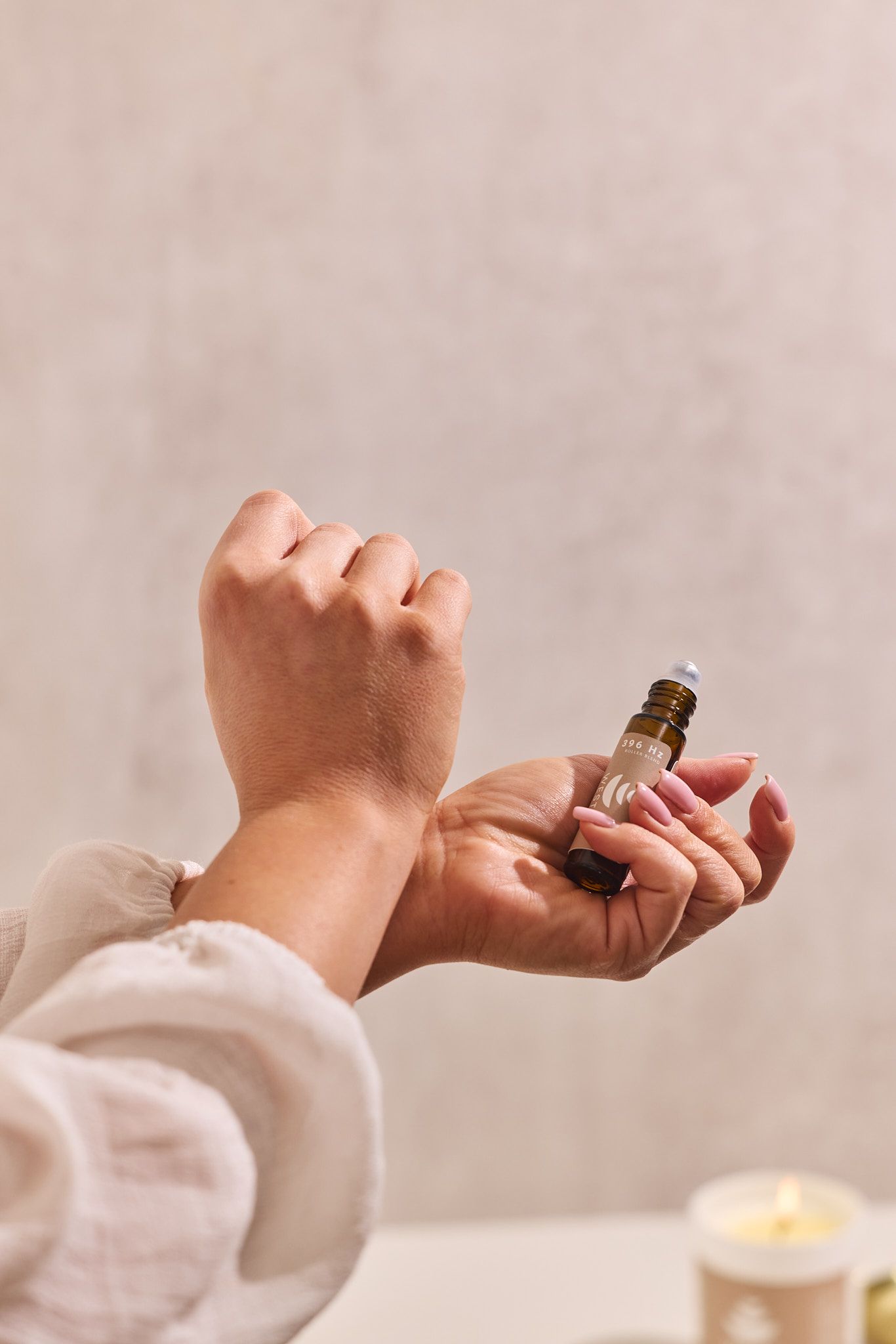 Hands holding a small bottle of essential oil or aromatherapy product with a candle on a light surface.